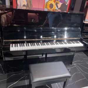 Glossy black J. Strauss & Sohn upright piano with a matching bench in a gallery setting, colorful paintings on the wall behind it.