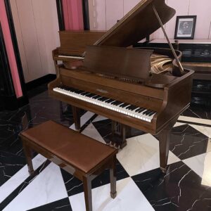 Brown grand piano with lid open and matching bench in a showroom-style room with pink walls and a black-and-white tiled floor.