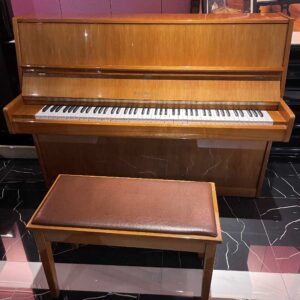 Wooden upright piano with matching cushioned bench in a showroom, keyboard visible beneath the lid.