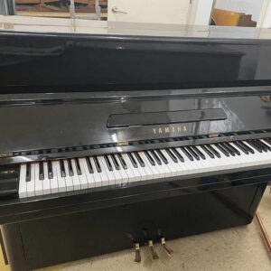 Black Yamaha upright piano in a workshop, keys exposed and three pedals visible